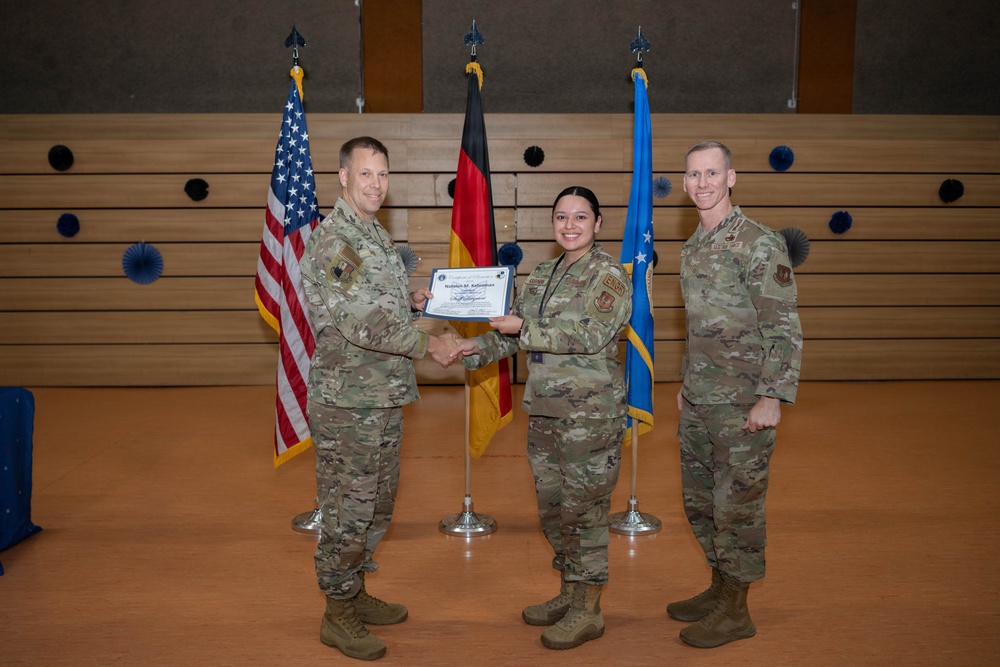 Three U.S. Air Force personnel in OCP (Operational Camouflage Pattern) uniforms are posed during a military awards or recognition ceremony. A certificate is being presented to a female service member by a senior officer while a third service member stands at attention to the right. The ceremony appears to be taking place in a formal venue with U.S., German, and U.S. Air Force flags displayed in the background.