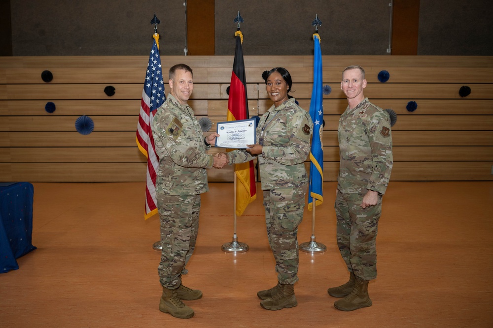 Three military personnel in OCP (Operational Camouflage Pattern) uniforms are posed during a formal award or recognition ceremony. A certificate is being presented to a female service member by a male colleague, while a third service member stands to the right. The setting appears to be a military installation, likely in Germany based on the visible German flag.