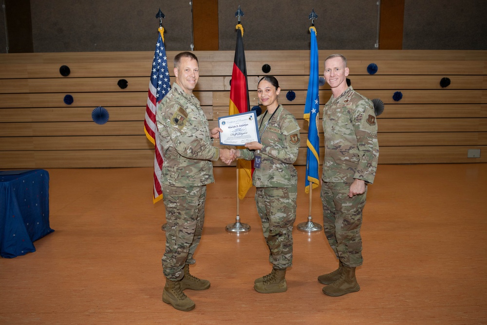 Three U.S. Air Force personnel in OCP (Operational Camouflage Pattern) uniforms are posed during a military promotion or award ceremony. A certificate is being presented to a female service member by a senior officer while a third service member stands to the right. The ceremony appears to take place in a formal venue with U.S., German, and U.S. Air Force flags displayed in the background.