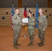 Three U.S. Air Force personnel in OCP (Operational Camouflage Pattern) uniforms are posed during a military promotion or award ceremony. A certificate is being presented to a female service member by a senior officer while a third service member stands to the right. The ceremony appears to take place in a formal venue with U.S., German, and U.S. Air Force flags displayed in the background.