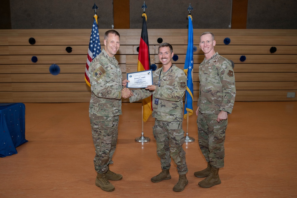 Three military personnel in U.S. Air Force OCP (Operational Camouflage Pattern) uniforms are posed for a formal award or recognition ceremony. The center individual is receiving a certificate from the person on the left while a third service member stands to the right. The setting appears to be an indoor ceremonial hall with U.S., German, and U.S. Air Force flags displayed in the background.