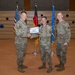 Three military personnel in U.S. Air Force OCP (Operational Camouflage Pattern) uniforms are posed for a formal award or recognition ceremony. The center individual is receiving a certificate from the person on the left while a third service member stands to the right. The setting appears to be an indoor ceremonial hall with U.S., German, and U.S. Air Force flags displayed in the background.