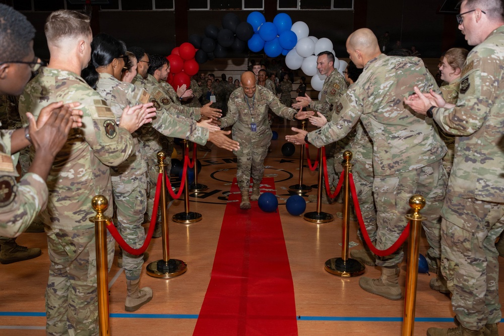 A military ceremony taking place inside a gymnasium or indoor sports facility, where a senior service member walks down a red carpet through a gauntlet of fellow uniformed personnel offering handshakes and applause. The event appears to be a formal recognition or farewell ceremony, decorated with red, white, blue, and black balloons. All personnel are wearing OCP (Operational Camouflage Pattern) uniforms consistent with U.S. Air Force or Army dress.