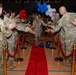A military ceremony taking place inside a gymnasium or indoor sports facility, where a senior service member walks down a red carpet through a gauntlet of fellow uniformed personnel offering handshakes and applause. The event appears to be a formal recognition or farewell ceremony, decorated with red, white, blue, and black balloons. All personnel are wearing OCP (Operational Camouflage Pattern) uniforms consistent with U.S. Air Force or Army dress.