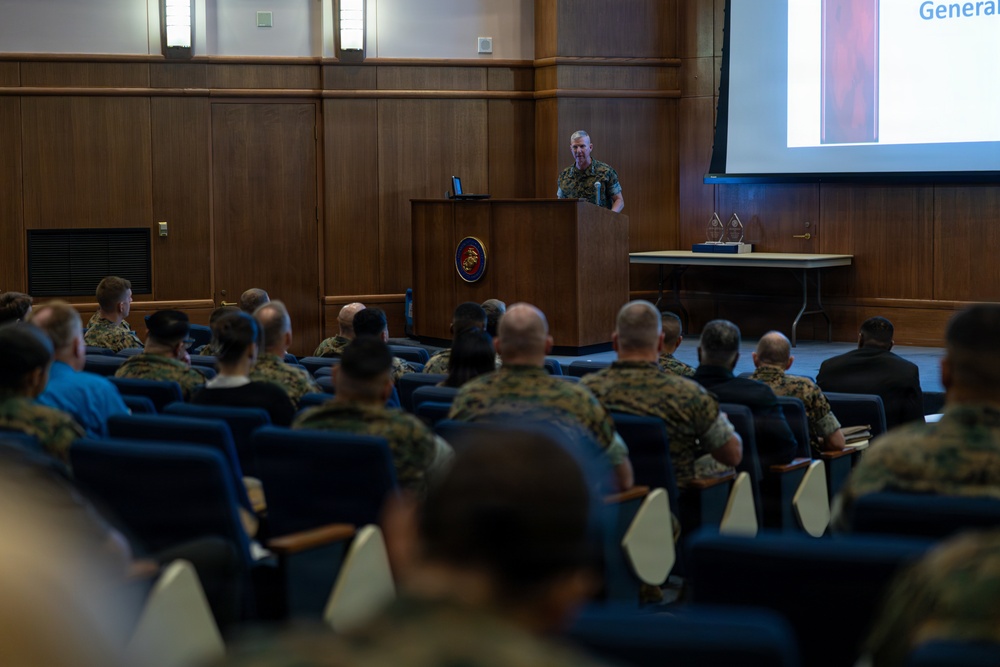 A military briefing or presentation is taking place in a formal auditorium setting. A uniformed officer stands at a wooden podium bearing what appears to be a U.S. Marine Corps emblem, addressing an audience of seated military personnel. A projection screen to the right displays a partially visible slide with the word 'General' visible.