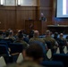 A military briefing or presentation is taking place in a formal auditorium setting. A uniformed officer stands at a wooden podium bearing what appears to be a U.S. Marine Corps emblem, addressing an audience of seated military personnel. A projection screen to the right displays a partially visible slide with the word 'General' visible.