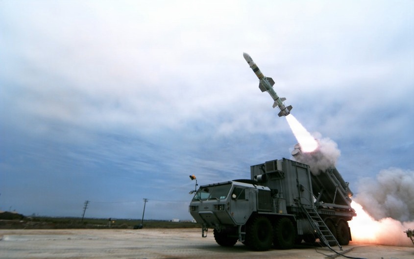 The image shows a missile being launched from a wheeled military vehicle on a flat, open terrain under an overcast sky. The missile is visibly ascending at an angle with a bright exhaust plume and smoke cloud at the launch point. The vehicle appears to be a multi-axle tactical truck fitted with a launcher module.