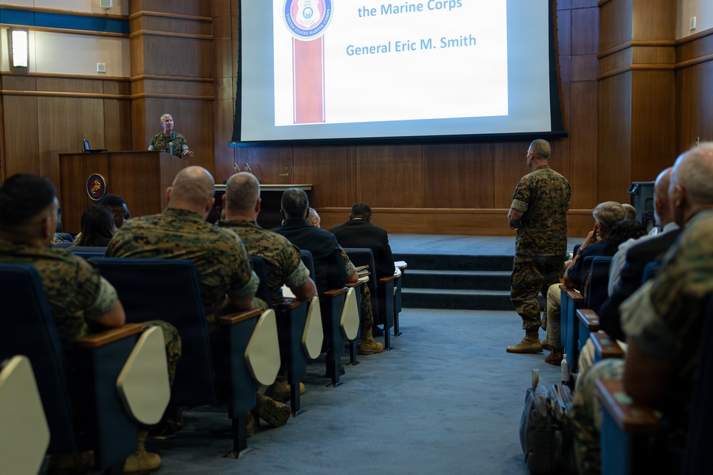 A military briefing or presentation is taking place in a formal auditorium setting, with a speaker at a podium addressing an audience of uniformed Marines and civilians. A large projection screen displays a slide referencing 'the Marine Corps' and 'General Eric M. Smith,' indicating this is an official U.S. Marine Corps presentation. The audience is seated in theater-style blue chairs, with one uniformed Marine standing in the aisle.