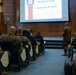 A military briefing or presentation is taking place in a formal auditorium setting, with a speaker at a podium addressing an audience of uniformed Marines and civilians. A large projection screen displays a slide referencing 'the Marine Corps' and 'General Eric M. Smith,' indicating this is an official U.S. Marine Corps presentation. The audience is seated in theater-style blue chairs, with one uniformed Marine standing in the aisle.