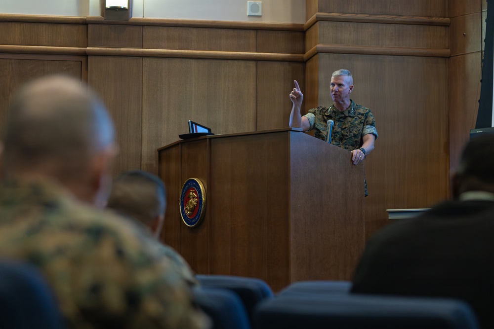 A U.S. Marine Corps officer in camouflage utility uniform is delivering a speech or briefing at a wooden podium bearing the official U.S. Marine Corps seal. He is gesturing with one finger raised, addressing an audience of military personnel seated in front of him. The setting appears to be a formal briefing room or auditorium within a Marine Corps facility.