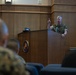 A U.S. Marine Corps officer in camouflage utility uniform is delivering a speech or briefing at a wooden podium bearing the official U.S. Marine Corps seal. He is gesturing with one finger raised, addressing an audience of military personnel seated in front of him. The setting appears to be a formal briefing room or auditorium within a Marine Corps facility.