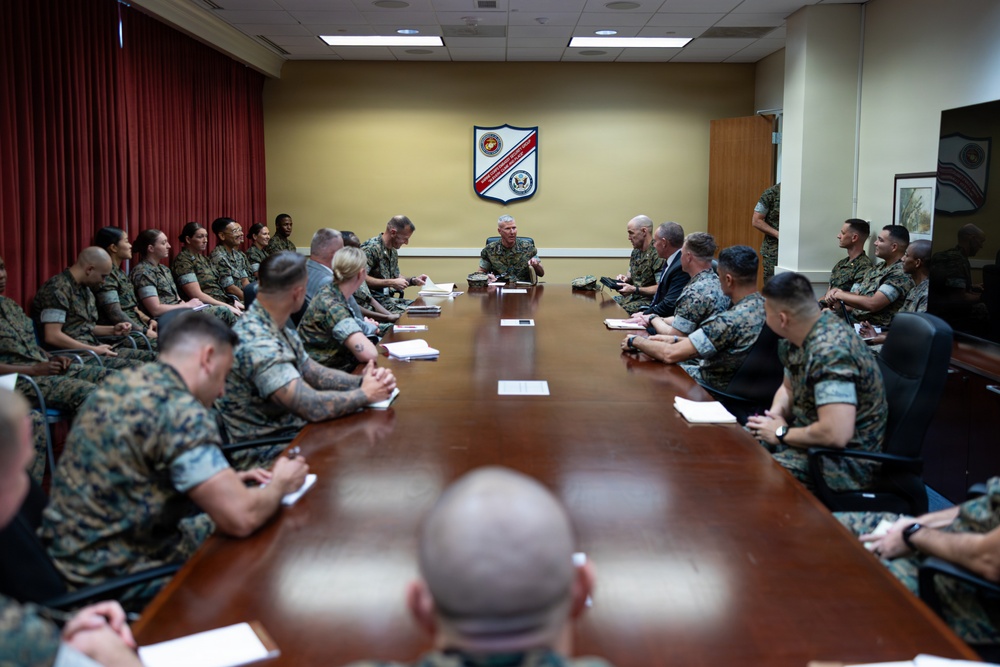 A large military briefing or meeting is taking place in a formal conference room, with numerous personnel in U.S. Marine Corps MARPAT camouflage uniforms seated around a long wooden conference table. A senior officer appears to be leading the discussion from the head of the table, with at least one civilian also present. A military unit shield/crest is prominently displayed on the wall behind the presiding officer.