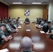 A large military briefing or meeting is taking place in a formal conference room, with numerous personnel in U.S. Marine Corps MARPAT camouflage uniforms seated around a long wooden conference table. A senior officer appears to be leading the discussion from the head of the table, with at least one civilian also present. A military unit shield/crest is prominently displayed on the wall behind the presiding officer.
