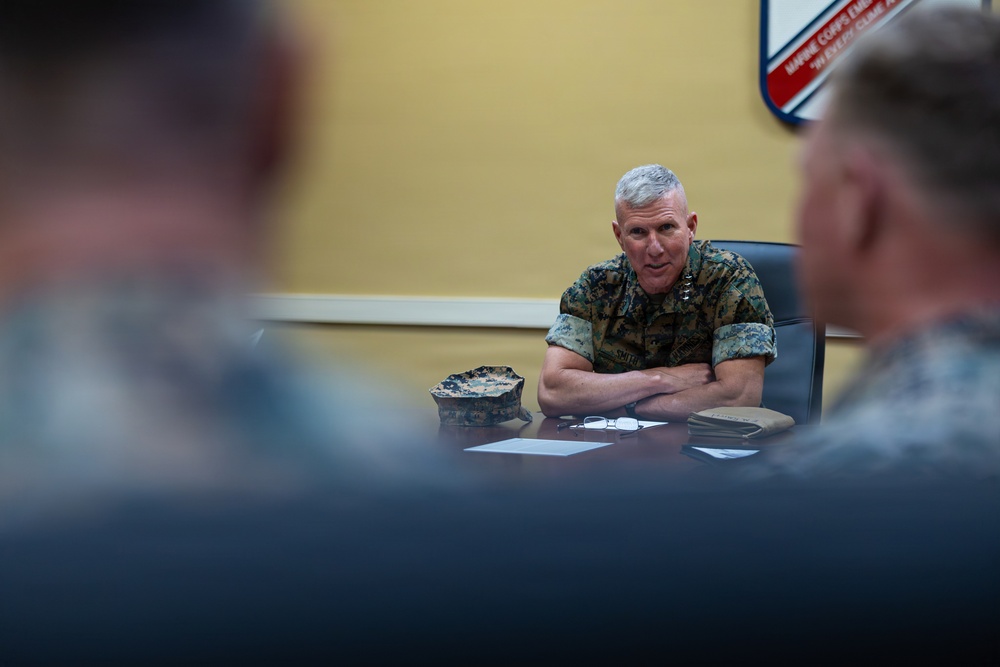 A senior military officer in U.S. Marine Corps camouflage uniform sits at a conference table with arms crossed, appearing to address or brief a group of personnel. A Marine Corps emblem or unit insignia is visible on the wall behind him. The foreground shows out-of-focus figures of other attendees seated across the table.