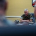 A senior military officer in U.S. Marine Corps camouflage uniform sits at a conference table with arms crossed, appearing to address or brief a group of personnel. A Marine Corps emblem or unit insignia is visible on the wall behind him. The foreground shows out-of-focus figures of other attendees seated across the table.