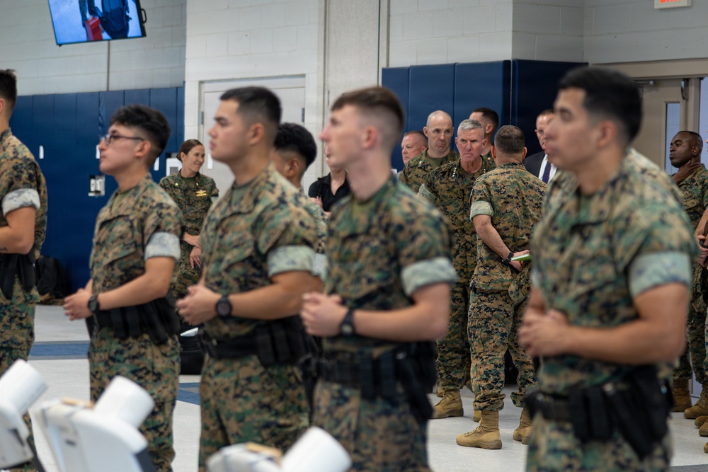 A group of U.S. Marines in MARPAT camouflage uniforms are assembled inside what appears to be a gymnasium or indoor facility. The personnel are standing at ease with hands clasped in front, suggesting a formal briefing or command visit. A senior officer, identifiable by rank insignia, is visible in the background engaging with other personnel.