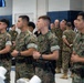 A group of U.S. Marines in MARPAT camouflage uniforms are assembled inside what appears to be a gymnasium or indoor facility. The personnel are standing at ease with hands clasped in front, suggesting a formal briefing or command visit. A senior officer, identifiable by rank insignia, is visible in the background engaging with other personnel.