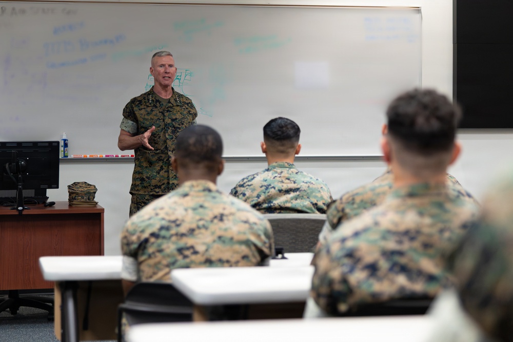 A senior military officer in U.S. Marine Corps MARPAT camouflage uniform is addressing a group of Marines in a classroom setting. The officer appears to be conducting a briefing or instructional session in front of a whiteboard with handwritten notes. Several Marines seated at desks are attentively listening with their backs to the camera.