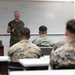 A senior military officer in U.S. Marine Corps MARPAT camouflage uniform is addressing a group of Marines in a classroom setting. The officer appears to be conducting a briefing or instructional session in front of a whiteboard with handwritten notes. Several Marines seated at desks are attentively listening with their backs to the camera.
