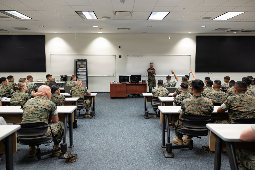 A military classroom setting shows a large group of uniformed personnel seated at individual desks facing an instructor at the front of the room. All individuals are wearing U.S. Marine Corps MARPAT camouflage uniforms. One student has their hand raised, indicating active participation in the instruction session.