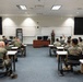 A military classroom setting shows a large group of uniformed personnel seated at individual desks facing an instructor at the front of the room. All individuals are wearing U.S. Marine Corps MARPAT camouflage uniforms. One student has their hand raised, indicating active participation in the instruction session.