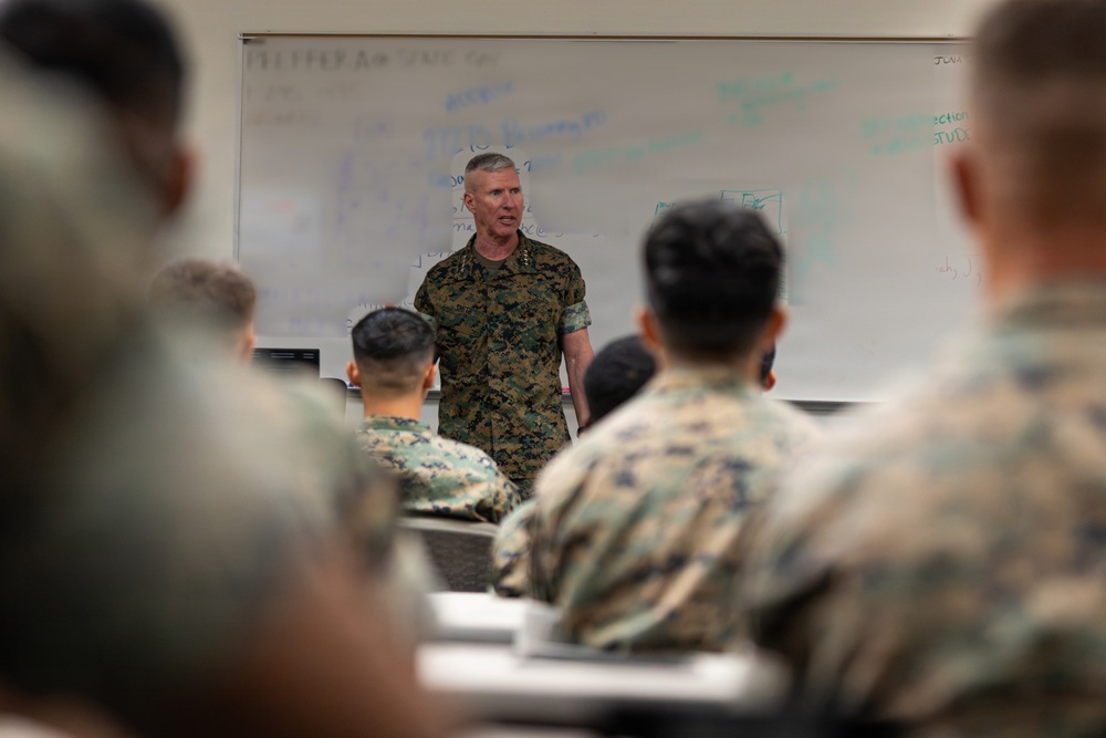 A senior military officer in MARPAT camouflage uniform stands at the front of a classroom addressing a group of seated military personnel. The setting appears to be a formal military instruction or briefing environment, with a whiteboard covered in notes and diagrams visible behind the speaker. The audience members, also in camouflage uniforms, are seated at desks facing the instructor.