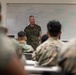 A senior military officer in MARPAT camouflage uniform stands at the front of a classroom addressing a group of seated military personnel. The setting appears to be a formal military instruction or briefing environment, with a whiteboard covered in notes and diagrams visible behind the speaker. The audience members, also in camouflage uniforms, are seated at desks facing the instructor.