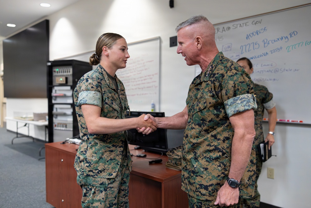 Two U.S. Marine Corps personnel in MARPAT woodland camouflage uniforms are shaking hands in what appears to be a military office or briefing room. A senior male officer with stars visible on his collar is greeting a female Marine. A whiteboard with written information is visible in the background along with a third person.