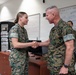 Two U.S. Marine Corps personnel in MARPAT woodland camouflage uniforms are shaking hands in what appears to be a military office or briefing room. A senior male officer with stars visible on his collar is greeting a female Marine. A whiteboard with written information is visible in the background along with a third person.
