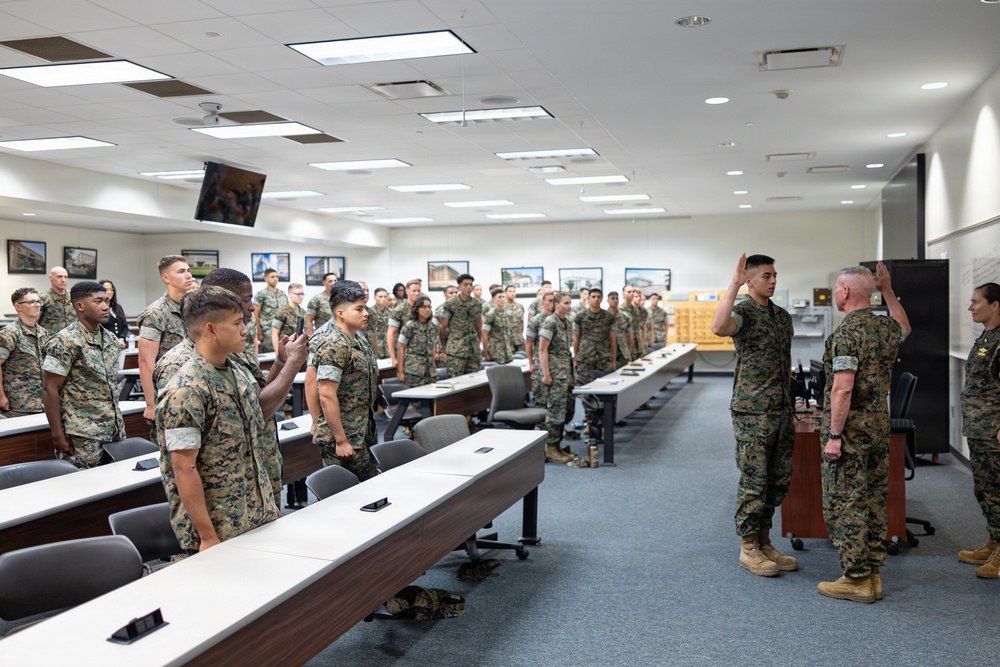 A group of U.S. Marines in woodland MARPAT camouflage uniforms are gathered in a large conference or briefing room. A ceremony appears to be taking place, with several individuals raising their right hands in what resembles an oath or reenlistment ceremony. A senior Marine on the right side appears to be administering the oath.