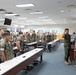 A group of U.S. Marines in woodland MARPAT camouflage uniforms are gathered in a large conference or briefing room. A ceremony appears to be taking place, with several individuals raising their right hands in what resembles an oath or reenlistment ceremony. A senior Marine on the right side appears to be administering the oath.