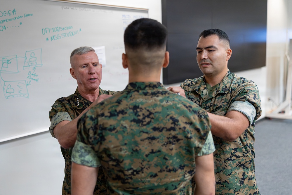 Three U.S. Marines in MARPAT camouflage uniforms are engaged in what appears to be a training or instructional interaction in a classroom setting. A senior officer on the left, identifiable by rank insignia on his collar, is speaking while both he and another Marine place hands on the shoulders of a third Marine facing away from the camera. A whiteboard with handwritten notes is visible in the background.