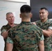 Three U.S. Marines in MARPAT camouflage uniforms are engaged in what appears to be a training or instructional interaction in a classroom setting. A senior officer on the left, identifiable by rank insignia on his collar, is speaking while both he and another Marine place hands on the shoulders of a third Marine facing away from the camera. A whiteboard with handwritten notes is visible in the background.