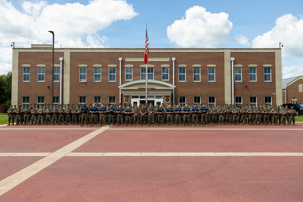A large group of military personnel, predominantly in camouflage uniforms with a smaller central group in blue shirts, pose for a formal group photograph in front of McMahon Hall, a three-story brick building on what appears to be a U.S. military installation. An American flag and a red pennant flag are visible on a flagpole centered in front of the building. The formation spans the full width of the brick plaza in front of the building.