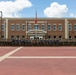 A large group of military personnel, predominantly in camouflage uniforms with a smaller central group in blue shirts, pose for a formal group photograph in front of McMahon Hall, a three-story brick building on what appears to be a U.S. military installation. An American flag and a red pennant flag are visible on a flagpole centered in front of the building. The formation spans the full width of the brick plaza in front of the building.