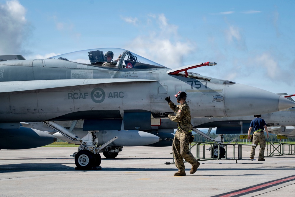 A Royal Canadian Air Force (RCAF) CF-18 Hornet fighter jet with tail number 757 is on an airfield tarmac. Ground crew personnel are conducting pre-flight or post-flight checks while a pilot sits in the cockpit. The aircraft bears the RCAF ARC roundel insignia and crew names 'Cplc Roy' and 'Cpt Ouellet' stenciled on the fuselage.