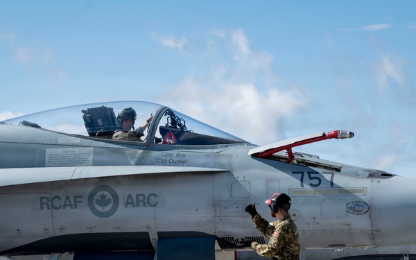 A Royal Canadian Air Force (RCAF) CF-18 Hornet fighter jet with tail number 757 is on an airfield tarmac. Ground crew personnel are conducting pre-flight or post-flight checks while a pilot sits in the cockpit. The aircraft bears the RCAF ARC roundel insignia and crew names 'Cplc Roy' and 'Cpt Ouellet' stenciled on the fuselage.