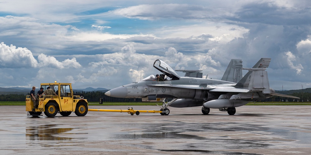 A Royal Canadian Air Force (RCAF) CF-18 Hornet fighter jet numbered 782 is being towed by a yellow aircraft tug (marked P-7) on a wet tarmac. Ground crew personnel are visible operating the tug and attending to the aircraft, whose canopy is open with a pilot or crew member seated inside. The scene appears to be at a Canadian military airbase under partly cloudy skies.