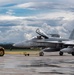 A Royal Canadian Air Force (RCAF) CF-18 Hornet fighter jet numbered 782 is being towed by a yellow aircraft tug (marked P-7) on a wet tarmac. Ground crew personnel are visible operating the tug and attending to the aircraft, whose canopy is open with a pilot or crew member seated inside. The scene appears to be at a Canadian military airbase under partly cloudy skies.
