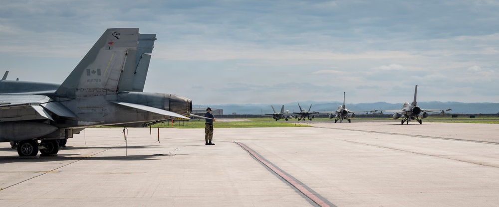 A military airfield scene showing a Canadian Armed Forces CF-18 Hornet in the foreground with its tail section prominently displaying a Canadian maple leaf flag insignia. Multiple fighter jets, including what appear to be F-16s and additional CF-18s, are taxiing on the runway in the background. A single ground crew member in military uniform stands on the tarmac, likely directing or observing aircraft movements.
