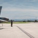 A military airfield scene showing a Canadian Armed Forces CF-18 Hornet in the foreground with its tail section prominently displaying a Canadian maple leaf flag insignia. Multiple fighter jets, including what appear to be F-16s and additional CF-18s, are taxiing on the runway in the background. A single ground crew member in military uniform stands on the tarmac, likely directing or observing aircraft movements.