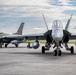 Two military fighter jets are parked on a concrete flight line apron at what appears to be a military air base. The foreground aircraft is an F/A-18 Hornet or Super Hornet viewed head-on, while the background aircraft is an F-16 Fighting Falcon. At least one ground crew member is visible crouching near the aircraft on the tarmac.