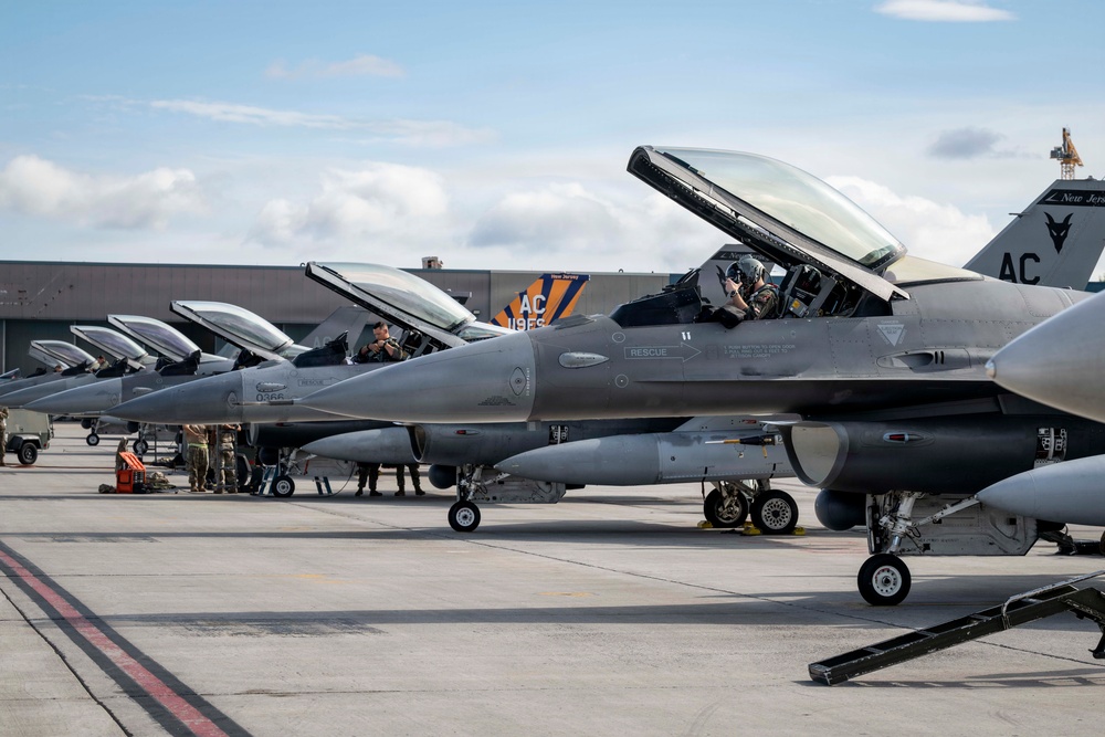 A line of F-16 Fighting Falcon jet fighters are parked on a flight line apron, with canopies open and pilots seated in cockpits. The aircraft bear 'AC' tail codes and markings consistent with the New Jersey Air National Guard's 177th Fighter Wing based at Atlantic City Air National Guard Base. Ground crew personnel are visible performing pre-flight or maintenance activities alongside the aircraft.