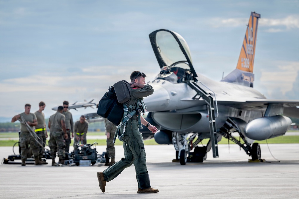 A military pilot in flight suit and harness walks across an airfield tarmac carrying a flight bag, with an F-16 Fighting Falcon jet fighter in the background. Ground crew personnel are visible working near the aircraft, which has its canopy open and an orange tail marking. The scene appears to depict pre-flight or post-flight operations at a military air base.