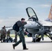 A military pilot in flight suit and harness walks across an airfield tarmac carrying a flight bag, with an F-16 Fighting Falcon jet fighter in the background. Ground crew personnel are visible working near the aircraft, which has its canopy open and an orange tail marking. The scene appears to depict pre-flight or post-flight operations at a military air base.