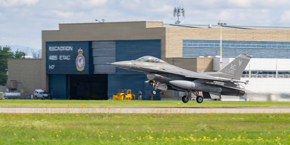 An F-16 Fighting Falcon jet fighter is captured during takeoff or landing on a runway, with its landing gear still deployed. The aircraft bears the tail code 'AC' and serial number '345', indicating it belongs to the New Jersey Air National Guard's 177th Fighter Wing. In the background, a Canadian military hangar marked 'ESCADRON 425 ETAC' is visible, suggesting this is a joint or exchange operation at a Canadian Forces base.