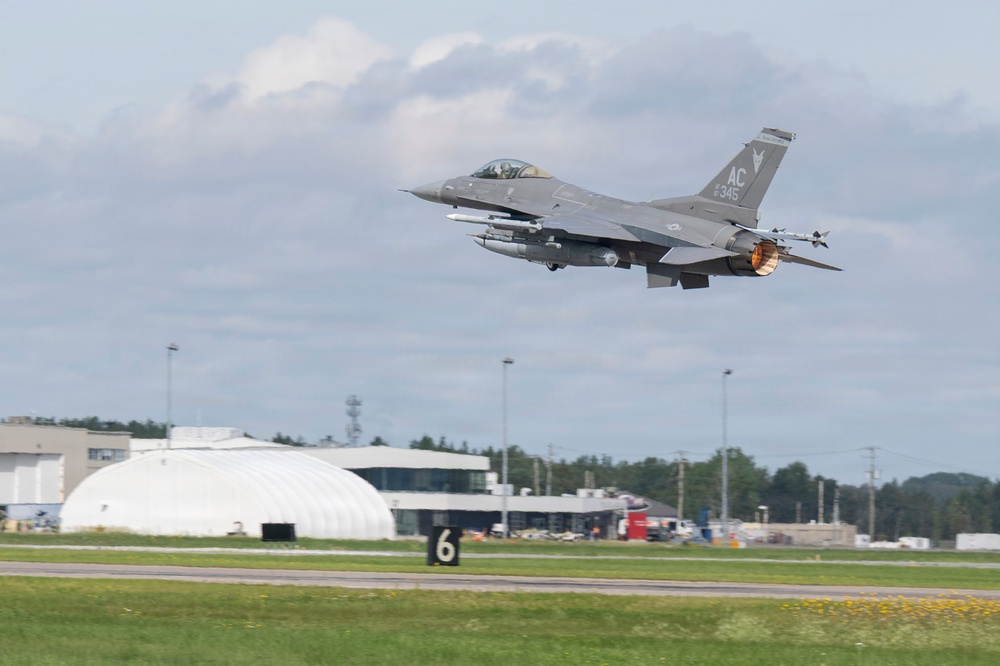 An F-16 Fighting Falcon jet fighter is captured just after takeoff from a military airfield, with afterburner visibly engaged. The aircraft bears tail markings 'AC' and serial number '345', consistent with the New Jersey Air National Guard's 177th Fighter Wing based at Atlantic City. The airfield infrastructure, including a white inflatable hangar and runway marker '6', is visible in the background.