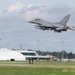 An F-16 Fighting Falcon jet fighter is captured just after takeoff from a military airfield, with afterburner visibly engaged. The aircraft bears tail markings 'AC' and serial number '345', consistent with the New Jersey Air National Guard's 177th Fighter Wing based at Atlantic City. The airfield infrastructure, including a white inflatable hangar and runway marker '6', is visible in the background.