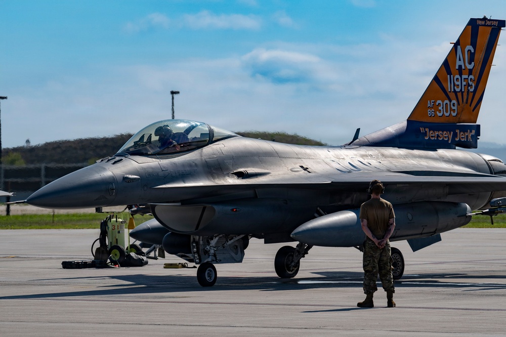 A U.S. Air Force F-16 Fighting Falcon assigned to the 119th Fighter Squadron, New Jersey Air National Guard, sits on a flight line tarmac with tail number AF 86-309, nicknamed 'Jersey Jerk.' A ground crew member stands at attention in front of the aircraft while a pilot is visible in the cockpit. The tail fin features the distinctive orange and blue New Jersey Air National Guard markings with 'AC 119FS' designation.