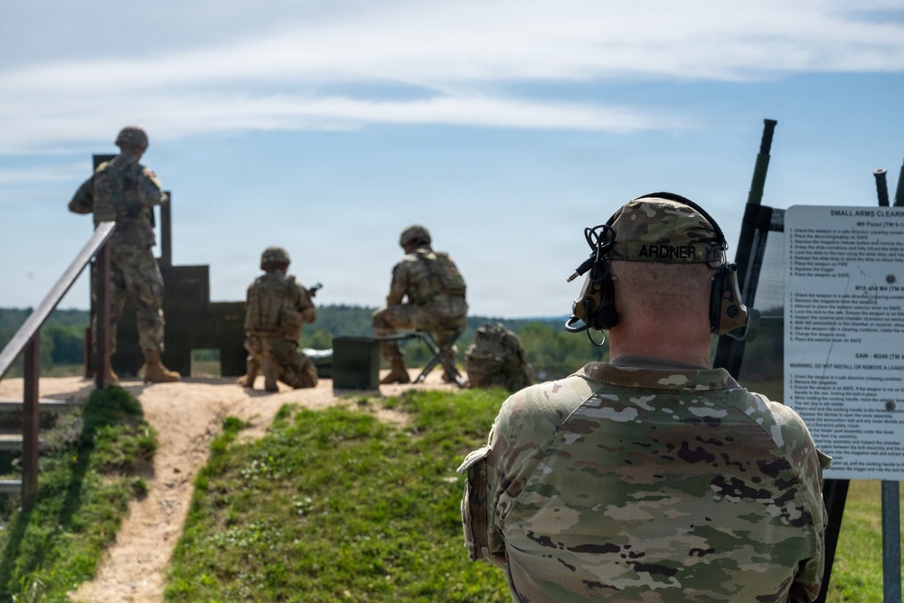 Military personnel are conducting a small arms range training exercise at an outdoor firing range. A soldier identified by name tape as 'ARDNER' is observing from a range control position, wearing tactical hearing protection. Multiple soldiers in OCP (Operational Camouflage Pattern) uniforms are visible downrange in various firing positions.